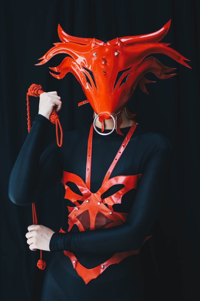 Eine weiße Person trägt eine rote Dämonenmaske mit Ring durch die Schnauze und einen roten Körperharness, der das dämonische Aussehen unterstreicht über enganliegender schwarzer Kleidung und hat eine rote Peitsche in der Hand.